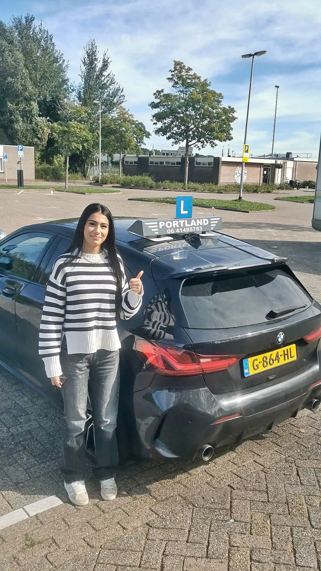 Een geslaagde leerling geeft een duim omhoog bij de lesauto met dakbord