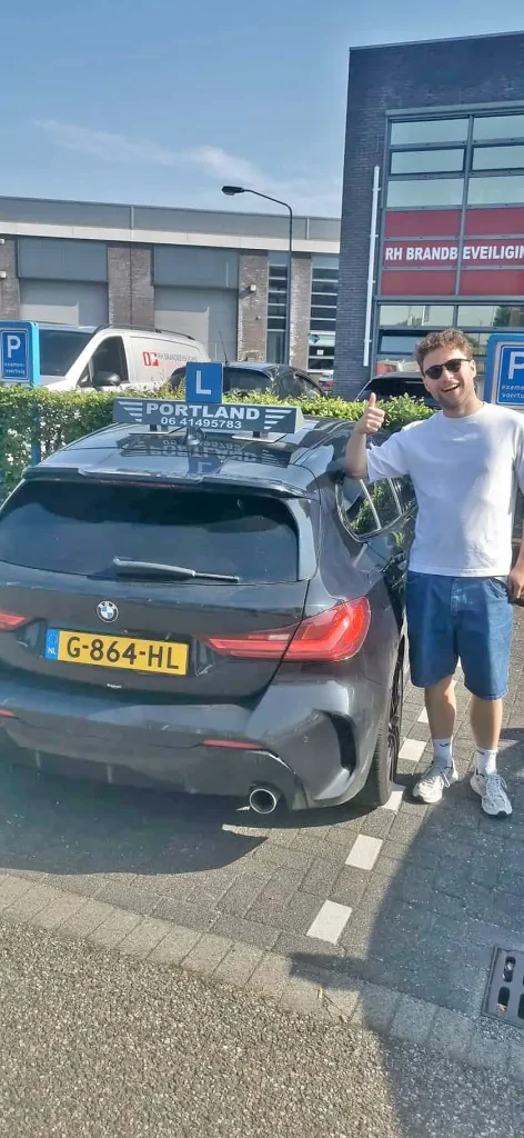 71e00076-e04b-44c4-92e2-9e5d7d5d9a79 Een blije leerling geeft een duim omhoog naast een lesauto van autorijschool Portland