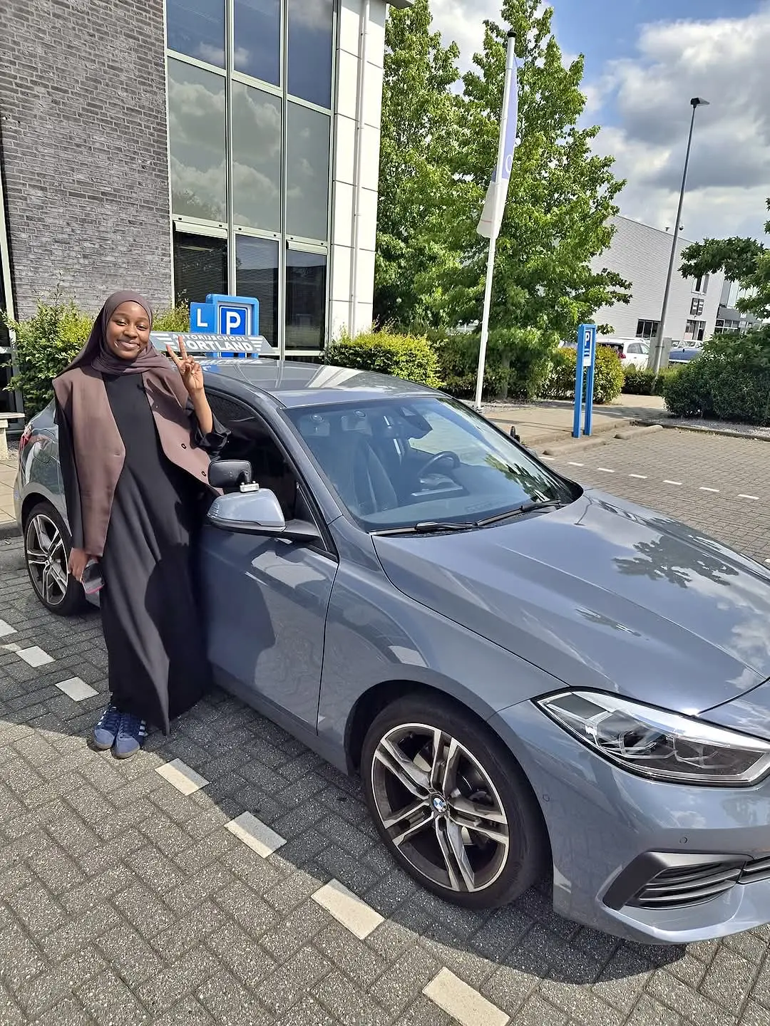 Een trotse leerling staat naast een grijze lesauto en maakt een V-teken