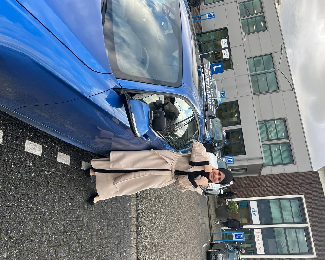 Geslaagde leerling staat naast een blauwe lesauto bij het CBR-gebouw