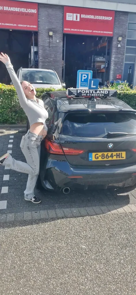 e308e0ef-d888-47fe-b7f5-46d4b072434f Een blije leerling staat juichend naast de lesauto na het behalen van haar rijbewijs